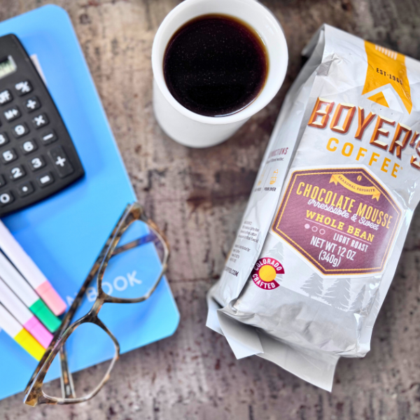 Boyer's Coffee package with a cup of coffee, glasses, and a notebook on a textured surface
