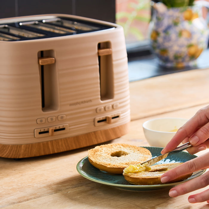 Person spreading butter on a bagel next to a beige toaster on a wooden table.