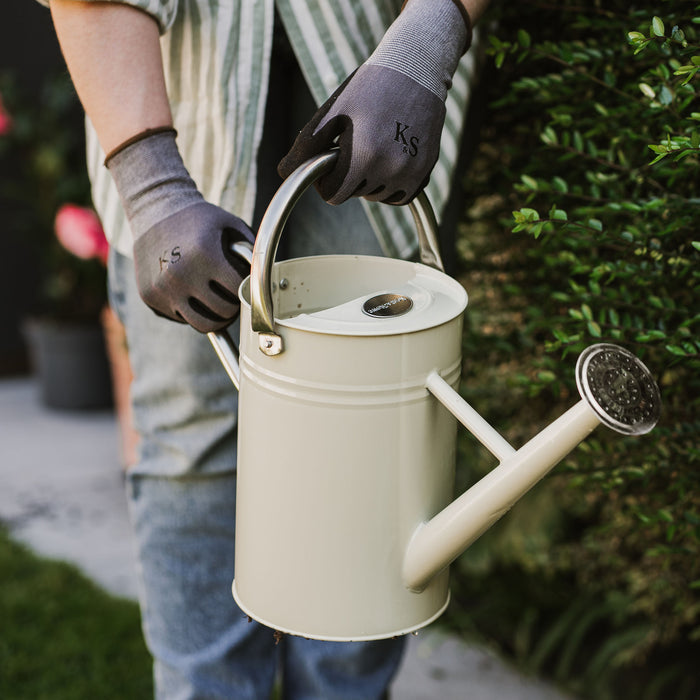 Person holding a beige watering can with a garden background, wearing seed and weed gloves
