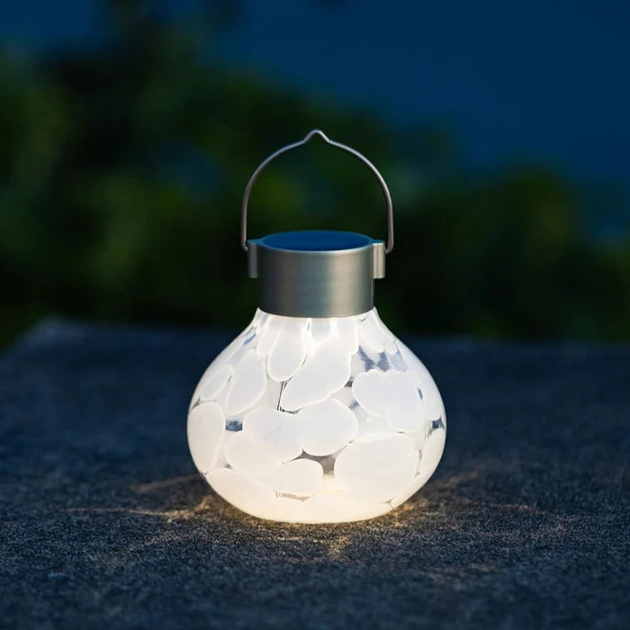 A glowing solar lantern with a white stone-look body and a metal handle, placed on a dark stone surface outdoors at night.